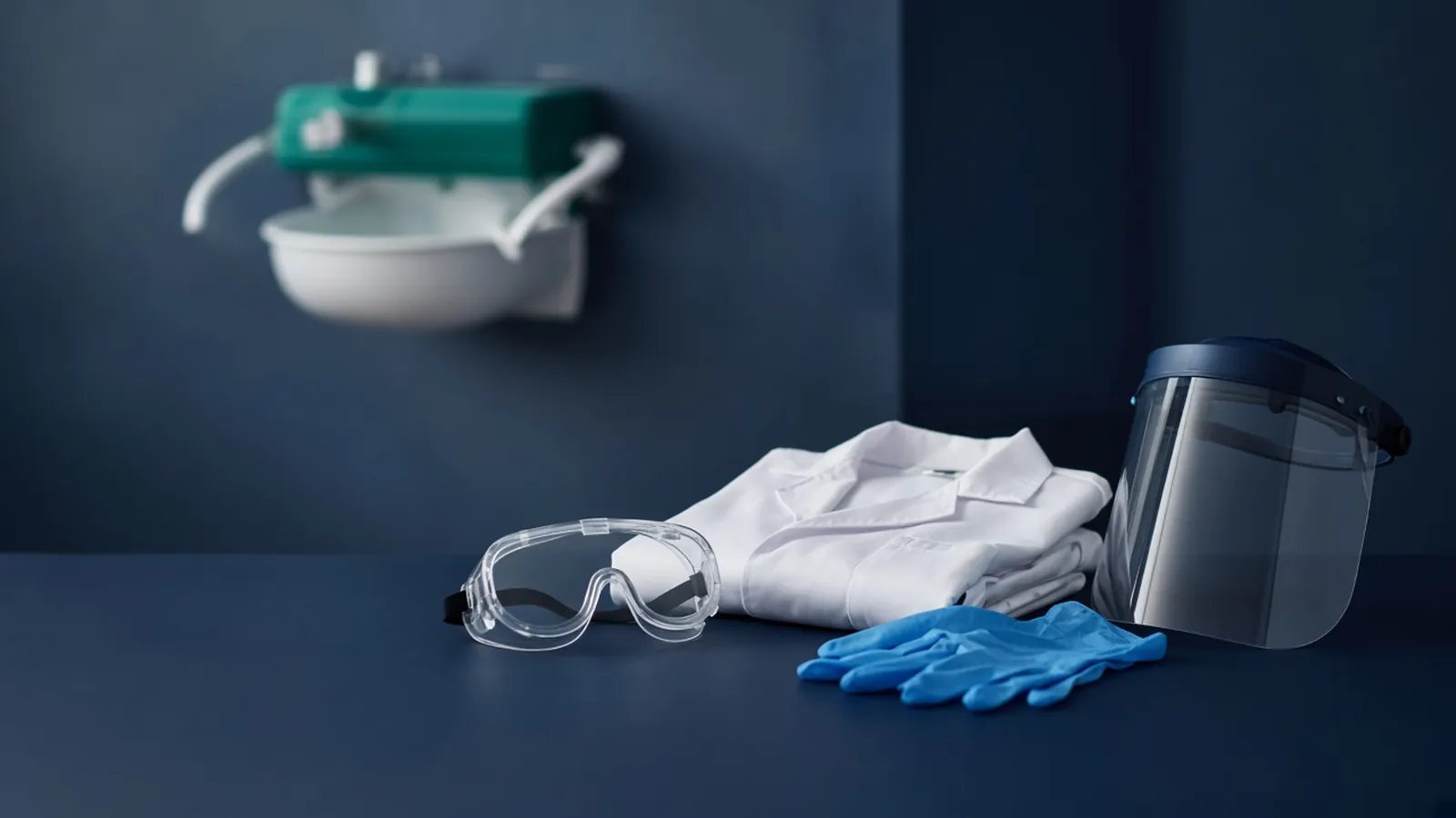 Safety goggles, lab coat, nitrile gloves and a face shield on a bench with an eyewash station behind